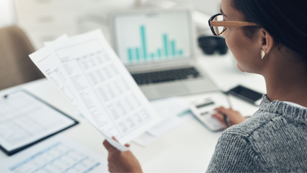 A woman in glasses reviews financial documents at her desk. She holds papers while analyzing data on a laptop with bar charts, conveying focus and analysis.