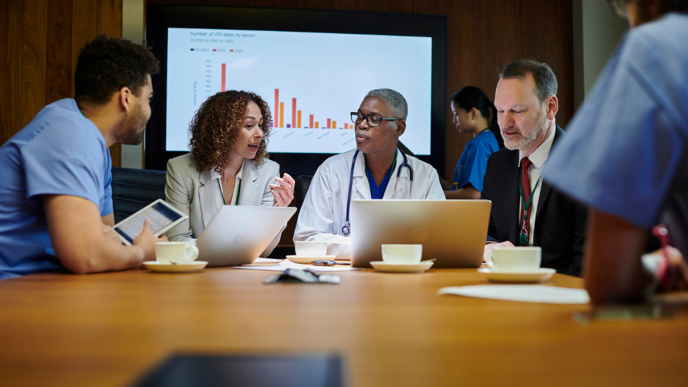 A group of professionals, including a doctor and business people, collaborate at a table with laptops. A screen shows a bar graph.