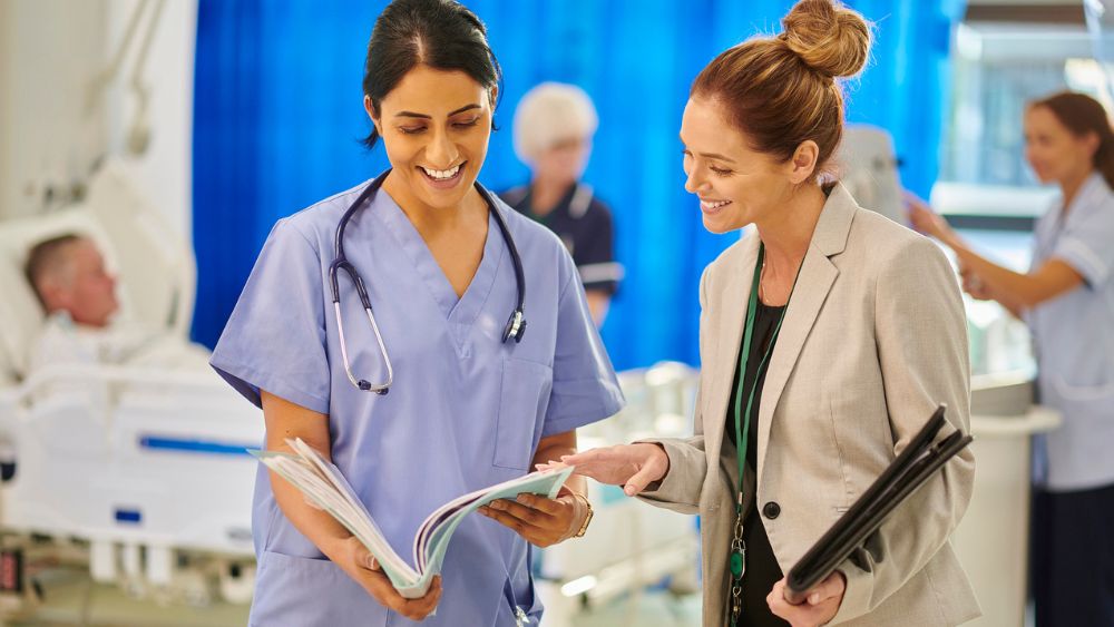 A nurse and a woman in a suit share a document, smiling in a busy hospital ward. Medical staff attend to patients in the background, conveying teamwork.