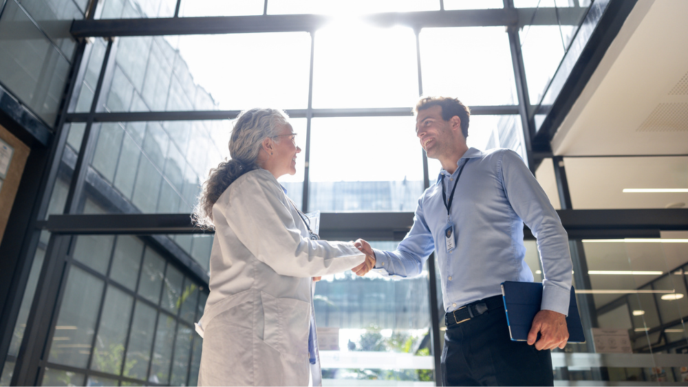 An older woman in a lab coat and a man in a blue shirt shake hands inside a brightly lit modern building, expressing professionalism and warmth.