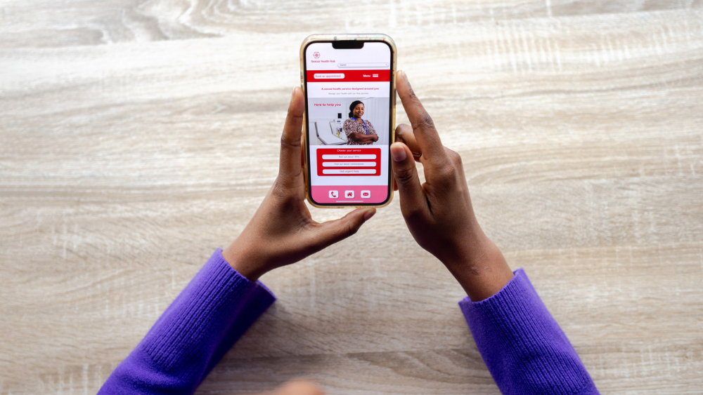 Hands holding a smartphone displaying a website with a red and white color scheme. The person wears a purple sweater, and the background is a wooden surface.